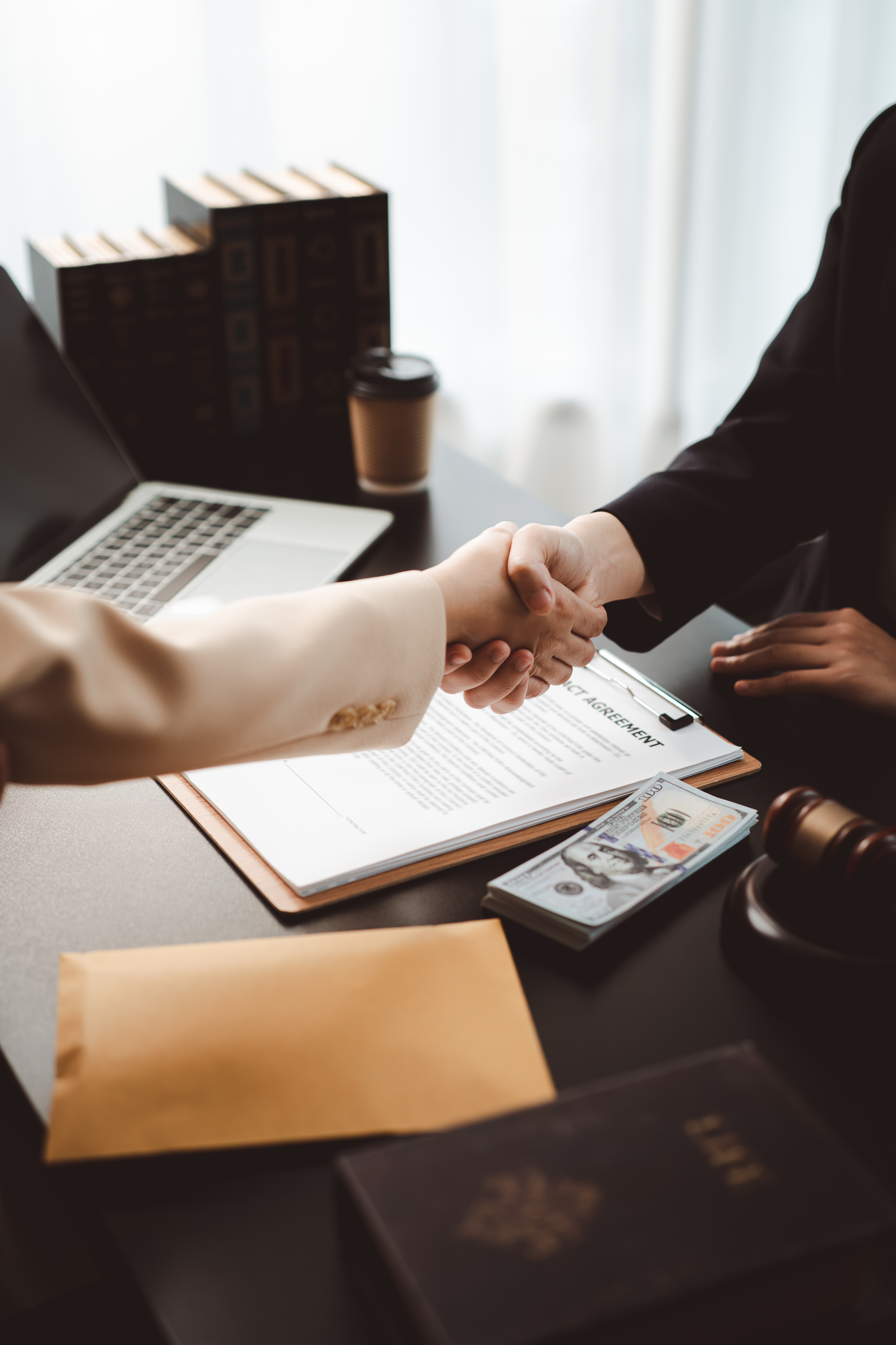Two business professionals shaking hands over a contract, money, and legal items on a desk, representing an agreement, a legal transaction, or a consultation with a lawyer
